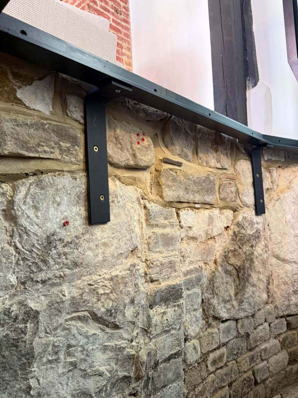 Powder coated black steel support brackets fixed into original stone wall beneath hinged glass floor, Burwash period property